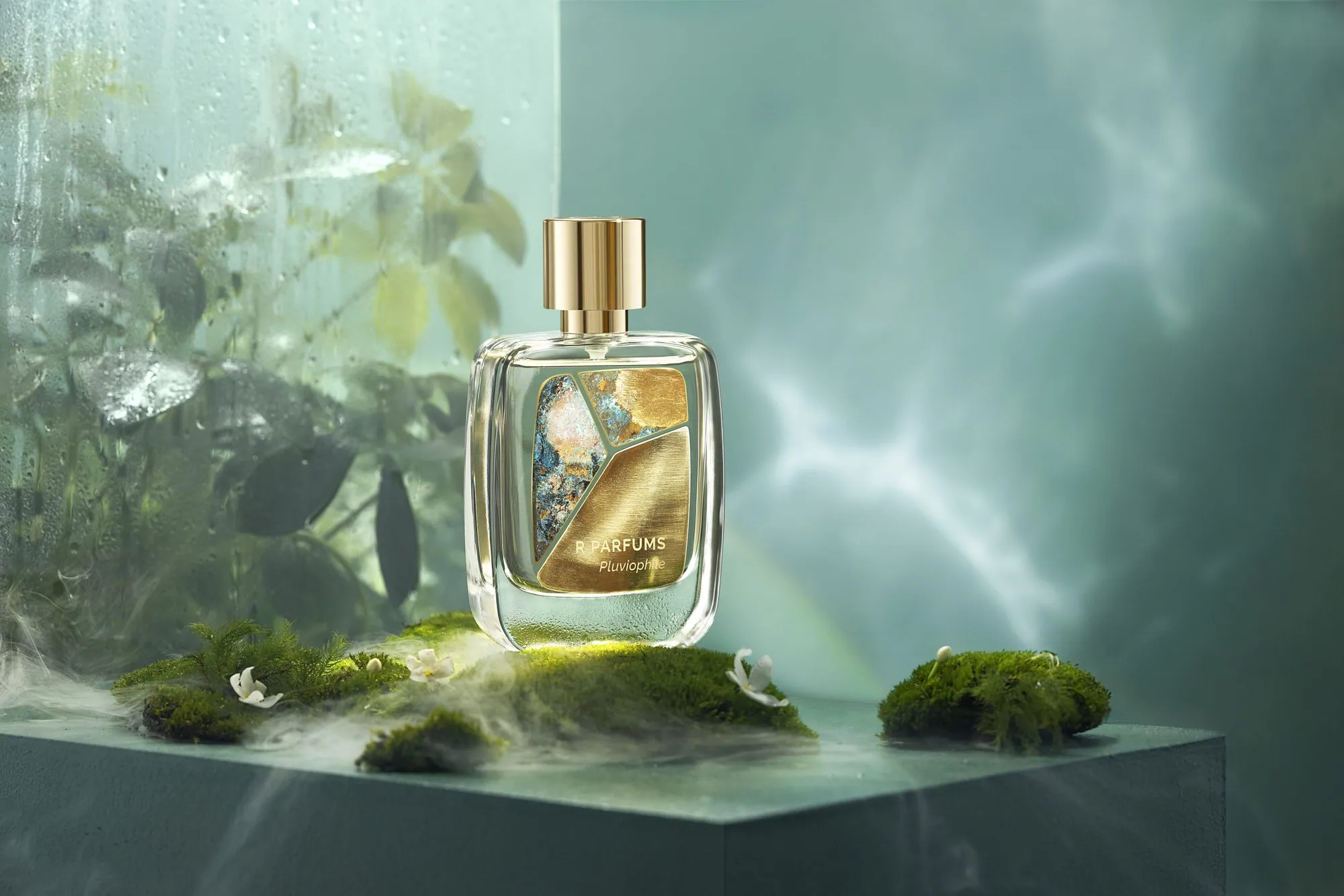 Chai Patina Pluviophile đặt trên rêu, giữa những bông hoa trắng nhỏ và làn sương trôi; phía sau là mưa trên ô kính và ánh sáng gợn như mặt nước
