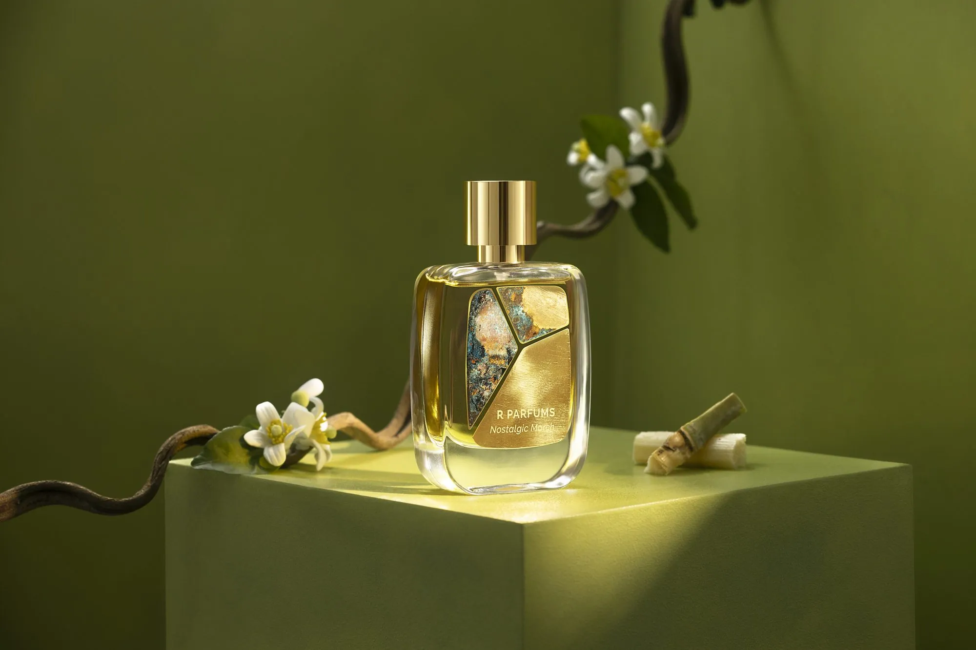 Product photograph, not of the house: Nostalgic March in the Patina bottle on a moss-green block, with a curving branch of white blossom and a length of sugarcane in slanting sunlight