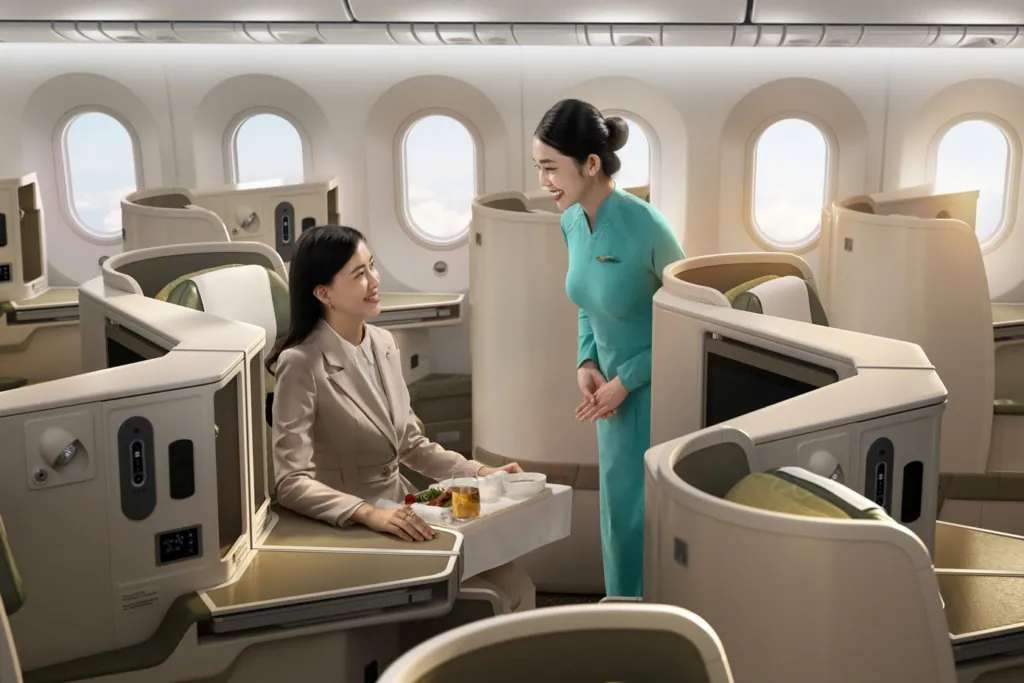 A flight attendant in teal áo dài chats with a business-class passenger over a tea tray