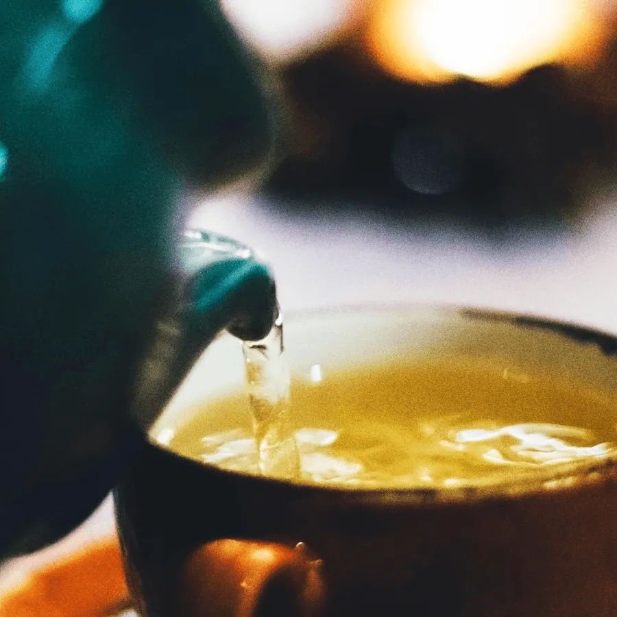 Gros plan sur un filet de thé vert doré versé de la théière dans la tasse, sur un fond de lumières chaudes et floues
