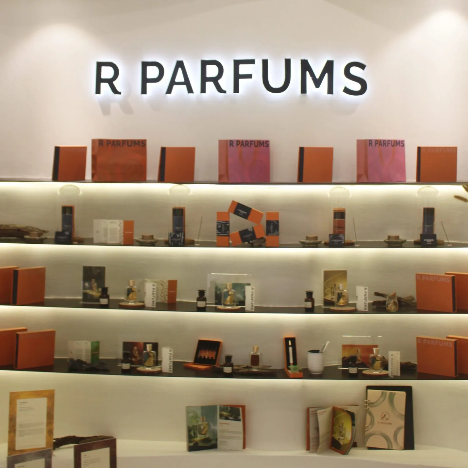 R PARFUMS 招牌下的陳列架：香水瓶、橘色與粉色禮盒、香水介紹卡，擺在一層層打亮的層架上。