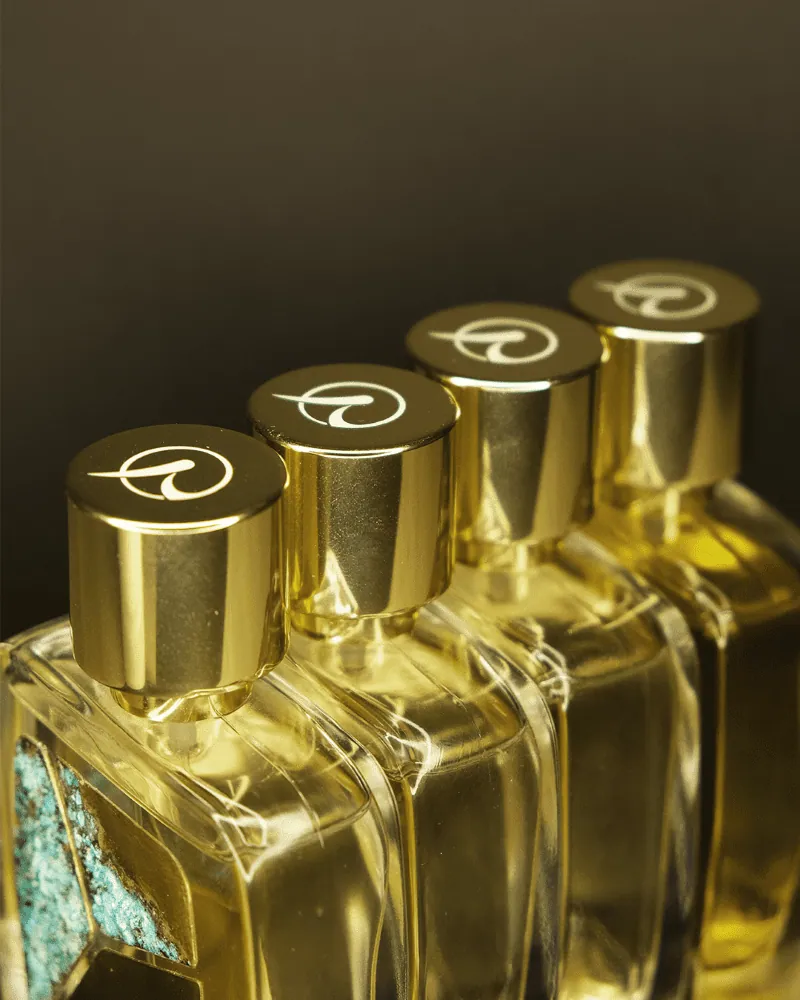 Four R Parfums bottles in a diagonal row, gold caps engraved with the R logo, a glimpse of turquoise patina below