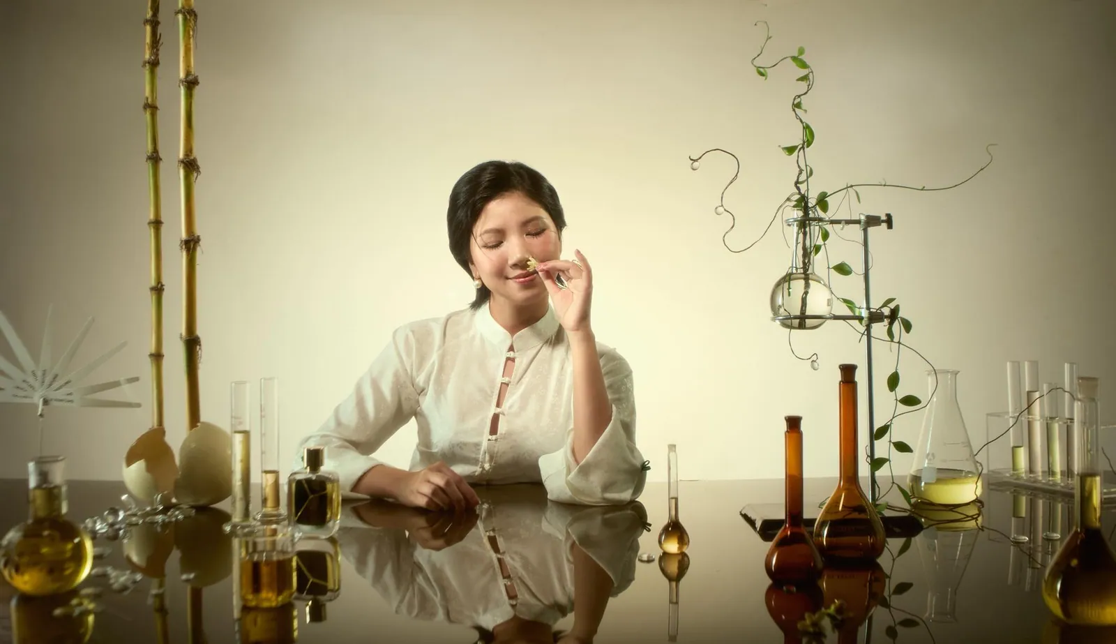 Perfumer Rei Nguyen eyes closed, smelling a bloom at a mirrored table of glass flasks and amber oils