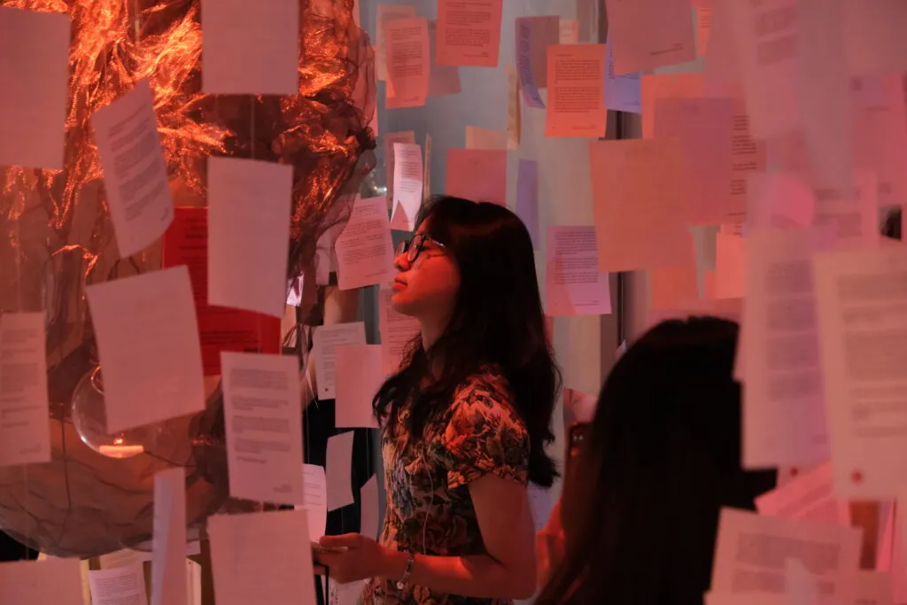 Des visiteurs debout parmi les pages suspendues, dans une lumière rose orangé, à l’exposition olfactive « Light and Shadow ».