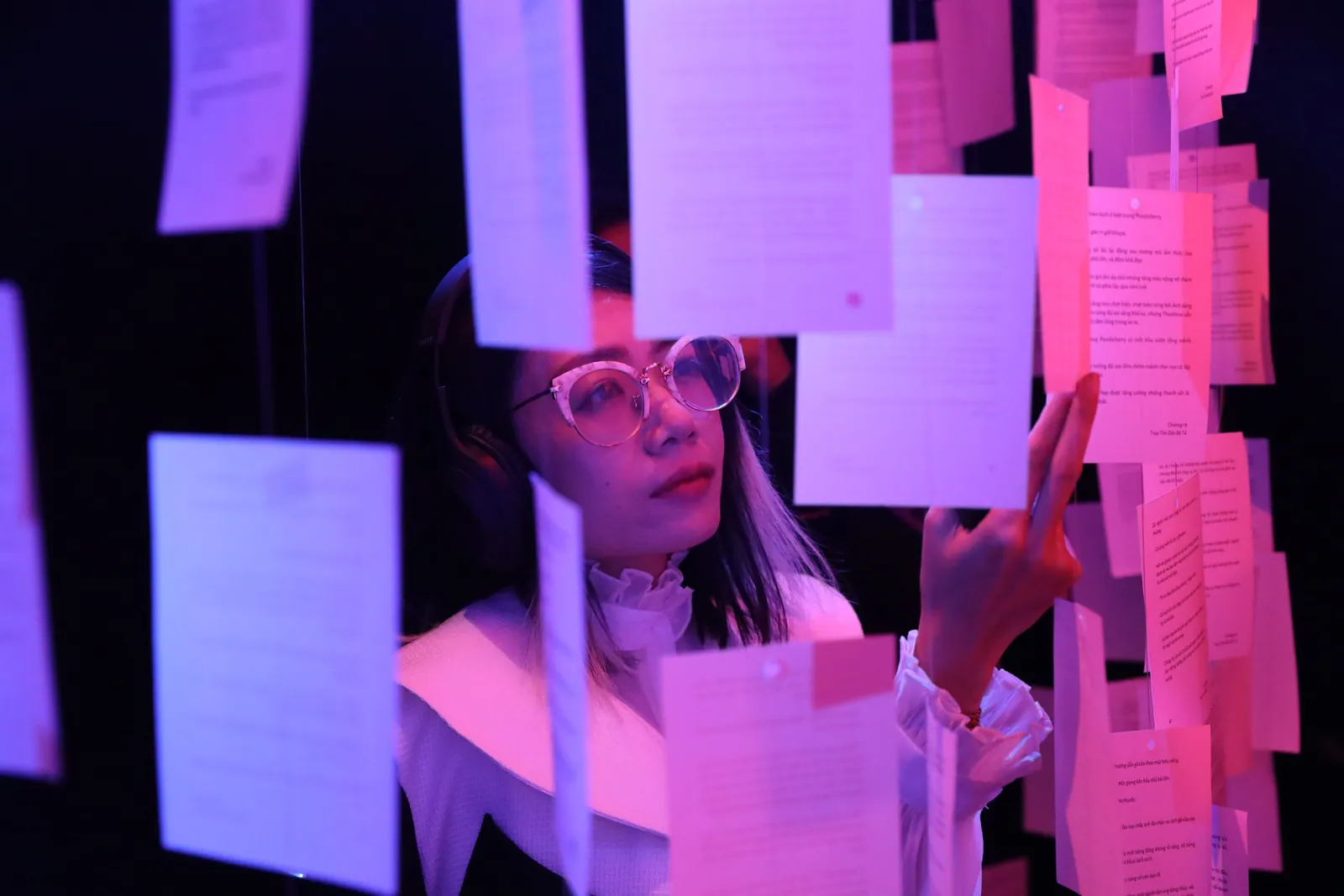 Une visiteuse à lunettes, casque sur les oreilles, touche une feuille suspendue pour lire l’histoire du parfum dans la lumière rose violacé