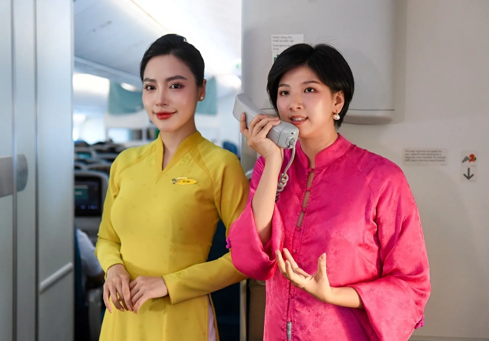 Rei Nguyen in pink speaks on the cabin interphone beside a flight attendant in a yellow áo dài