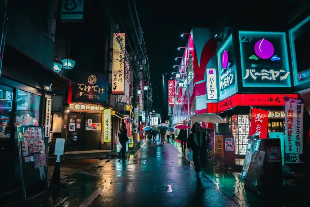 Hẻm Tokyo về đêm sau mưa, bảng hiệu neon phản chiếu mặt đường, người qua lại cầm ô