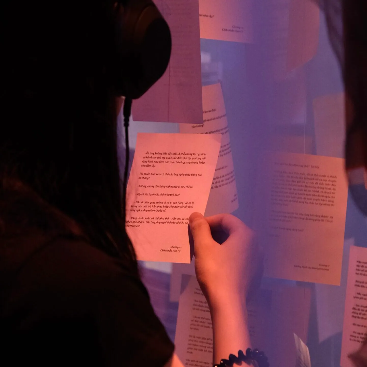 Casque sur les oreilles, un visiteur tient une page de récit suspendue et la lit, dans une lumière rose violacé, à « Light and Shadow ».
