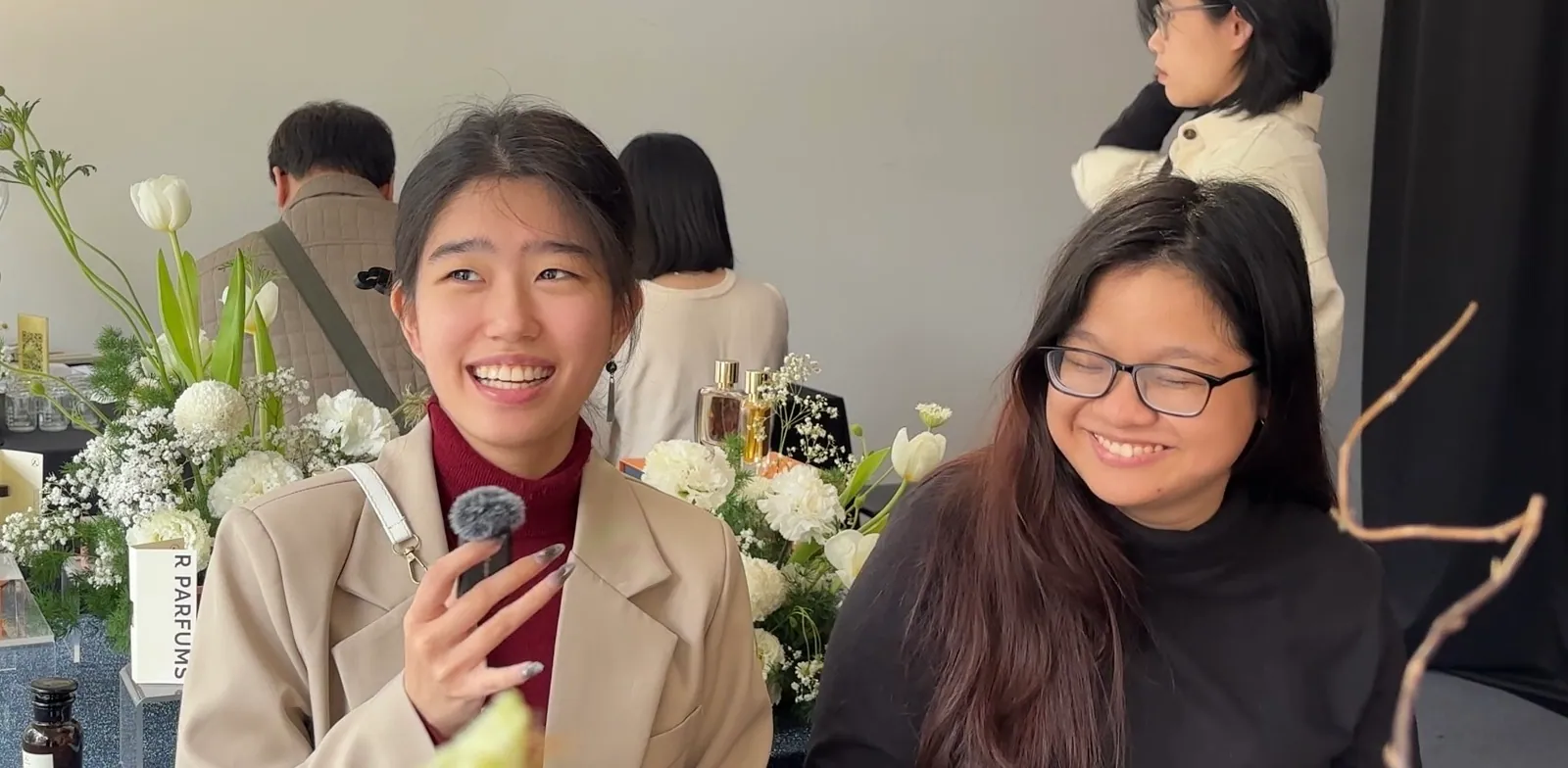 Two smiling visitors share their impressions beside a table of white flowers, perfume bottles and an R Parfums name plate
