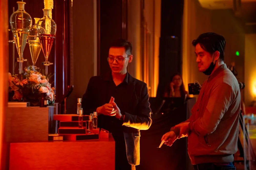 Une mouillette à la main, un visiteur parle avec la personne qui tient le stand R Parfums, sous une lumière orangée ; derrière, des entonnoirs de verre et des fleurs