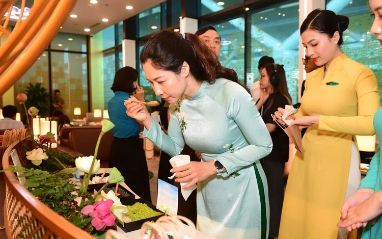 A guest in a pale teal áo dài samples food at a lotus and young-rice display beside an attendant in yellow