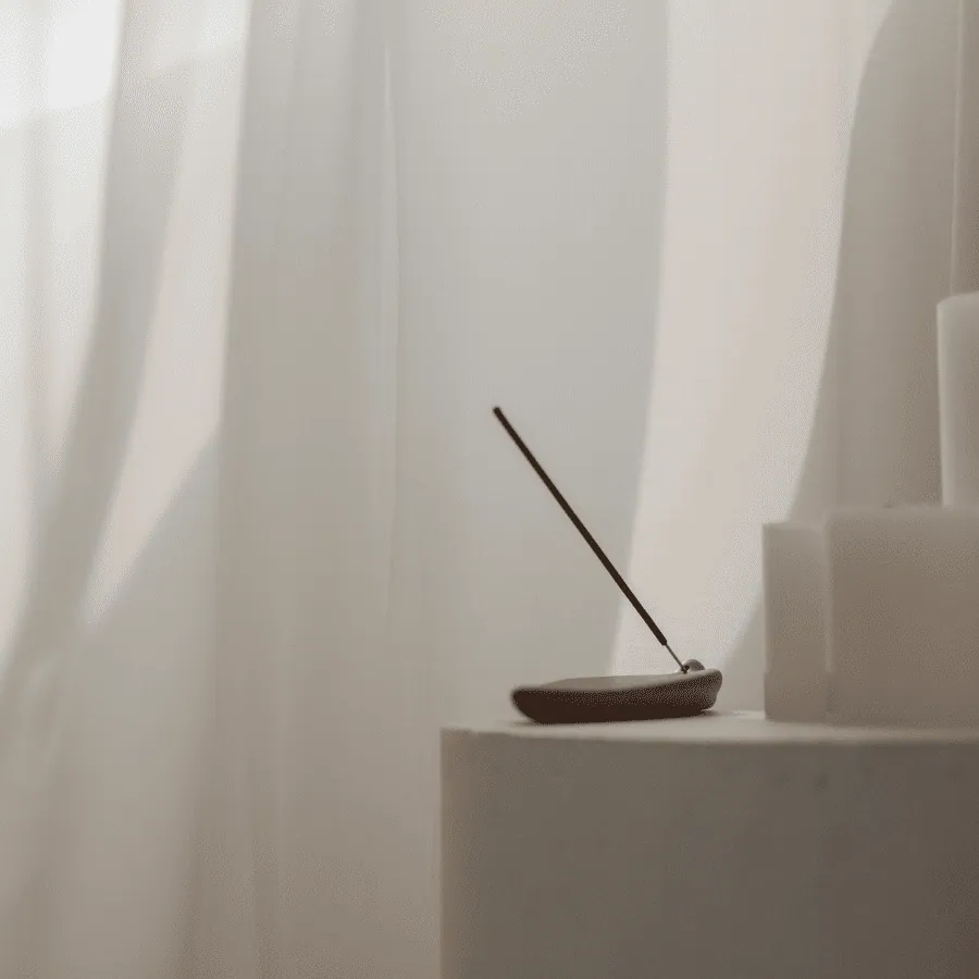 A single incense stick leaning from a dark stone holder beside white candles and sheer curtains