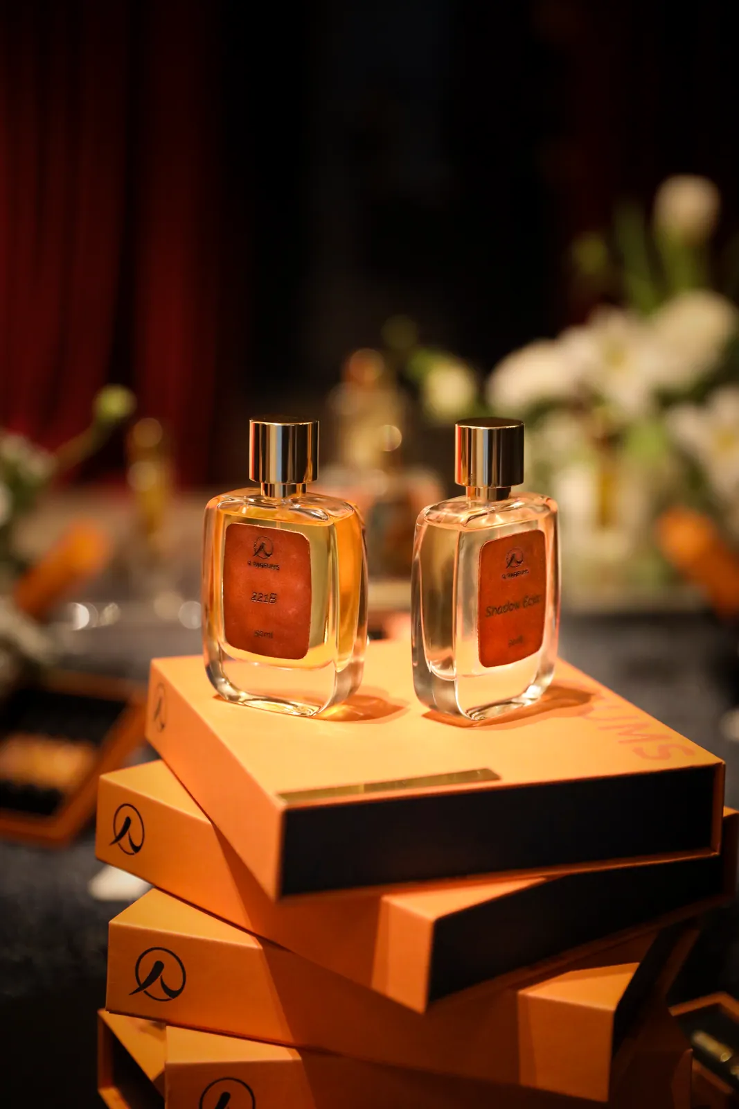 展覧会「Light and Shadow」の会場で、R Parfums のオレンジ色の箱を重ねた上に置かれた、茶色のレザーラベルの 221B と Shadow Éclat の 50 ml ボトル