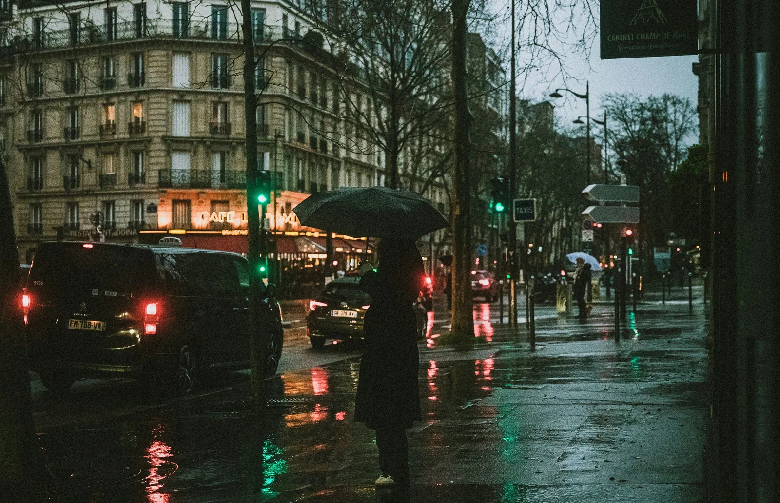 Người che ô đứng trên vỉa hè Paris ướt mưa lúc chạng vạng, đèn quán café và đèn xe phản chiếu