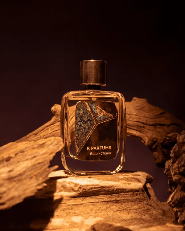 R Parfums の Béton Chaud のボトル。腐食させた真鍮のプレートに緑青が浮かび、温かな光のなか、流木のあいだに置かれている
