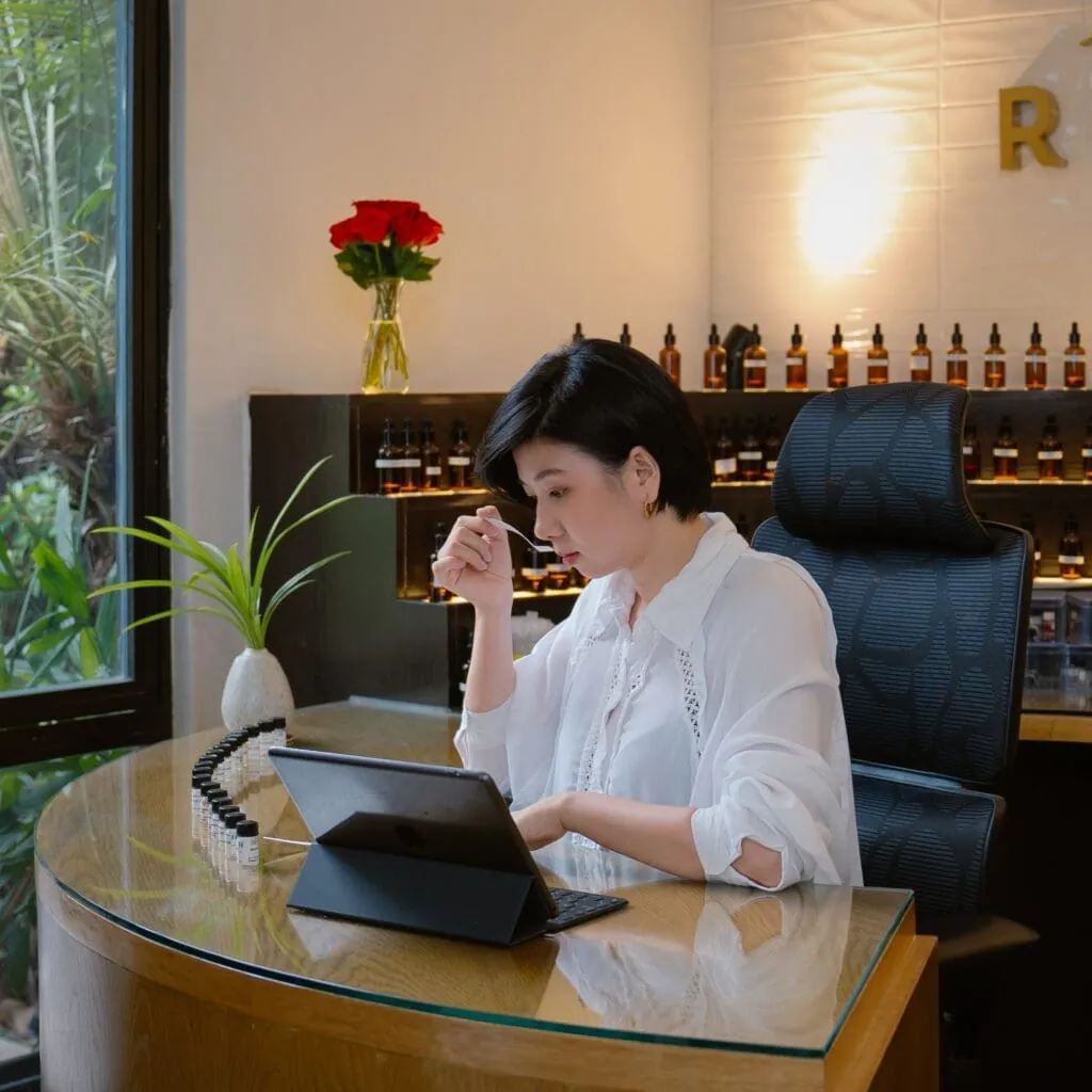 Người chế tác nước hoa ngửi que thử bên bàn làm việc, sau lưng là kệ lọ tinh dầu màu hổ phách