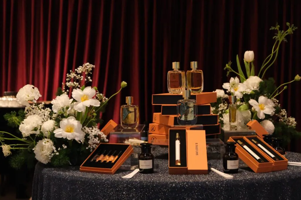 Table de présentation avec des flacons de parfum et des boîtes orange R Parfums près de fleurs blanches, devant un rideau de velours rouge, à « Light and Shadow »