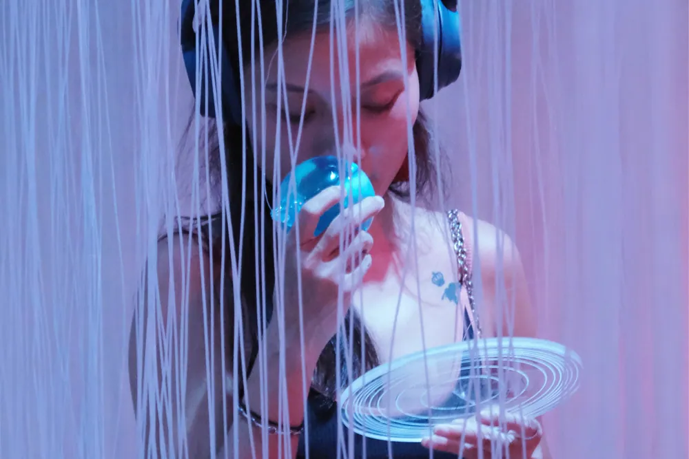 Une jeune femme portant un casque audio sent une sphère bleue, un disque transparent gravé de cercles à la main, devant un rideau de fils aux reflets violets et roses