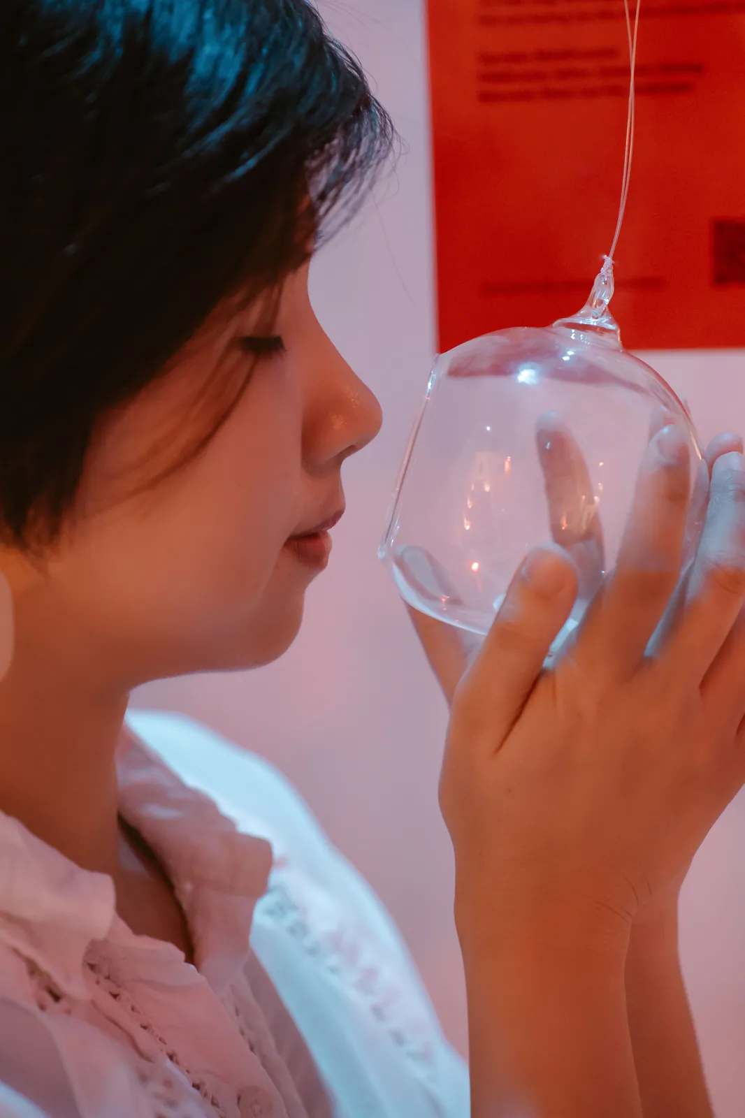 Une jeune femme aux cheveux courts, les yeux fermés, sent une sphère de verre suspendue à un fil, face à une œuvre d’art olfactif dans une exposition
