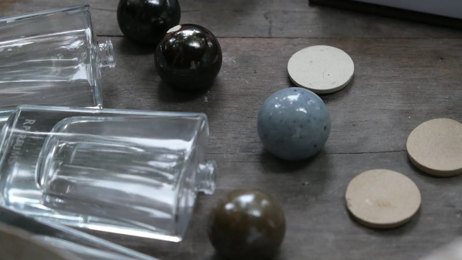 Un flacon de verre transparent à côté de bouchons en céramique terminés, à l’émail gris-bleu, brun et noir brillant, avec une breloque en céramique