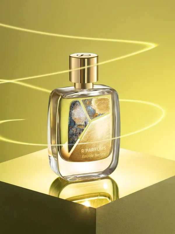 R Parfums Eau de Hajime 병, 손으로 마감한 황동 파티나 판, 굽이치는 빛줄기 사이 금빛 받침 위