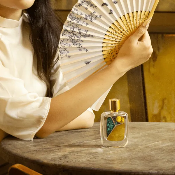 Une personne vêtue de blanc s’évente avec un éventail de papier aux fleurs peintes, à une vieille table en bois, près du flacon Nostalgic March
