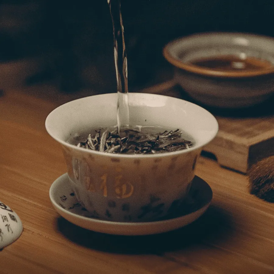 De l’eau versée dans un bol de porcelaine à couvercle qui contient des feuilles de thé sèches, sur une table en bois ; derrière, une tasse de thé déjà infusé