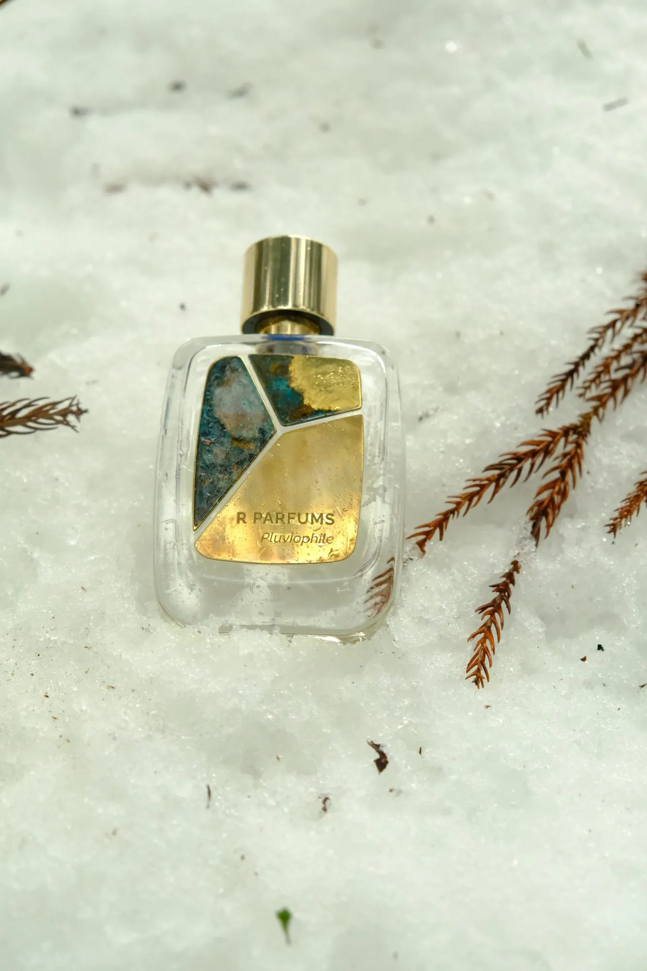Un flacon Pluviophile Patina dans la neige, de près : bouchon doré, un fragment de patine bleu-vert, un fragment mi-laiton mi-patine et un fragment de laiton brillant imprimé R PARFUMS Pluviophile ; à droite, des rameaux de conifère roux