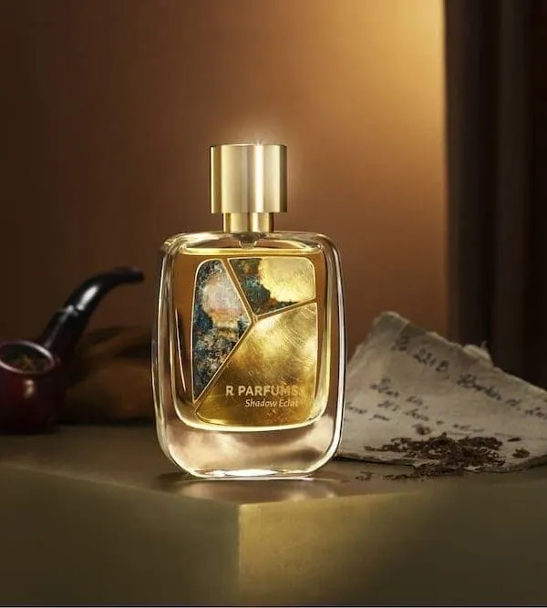 R Parfums Shadow Éclat 병과 황동 파티나 판, 나무 파이프와 낡은 편지 곁, 노란 불빛 아래