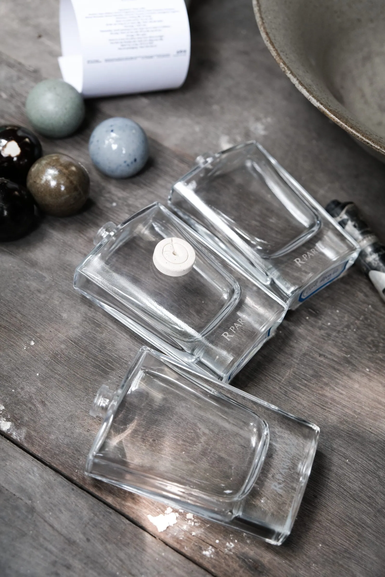 A square glass bottle with a ceramic disc at its neck, glazed spheres beside it