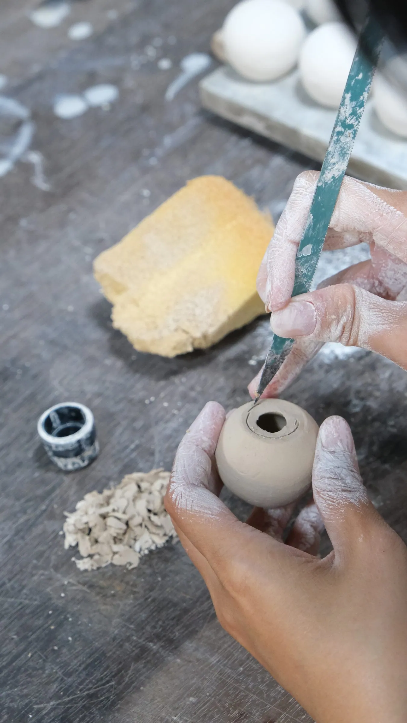 A green tool cutting the opening of a small clay sphere held in the hand, a sponge and crumbs of clay beside it