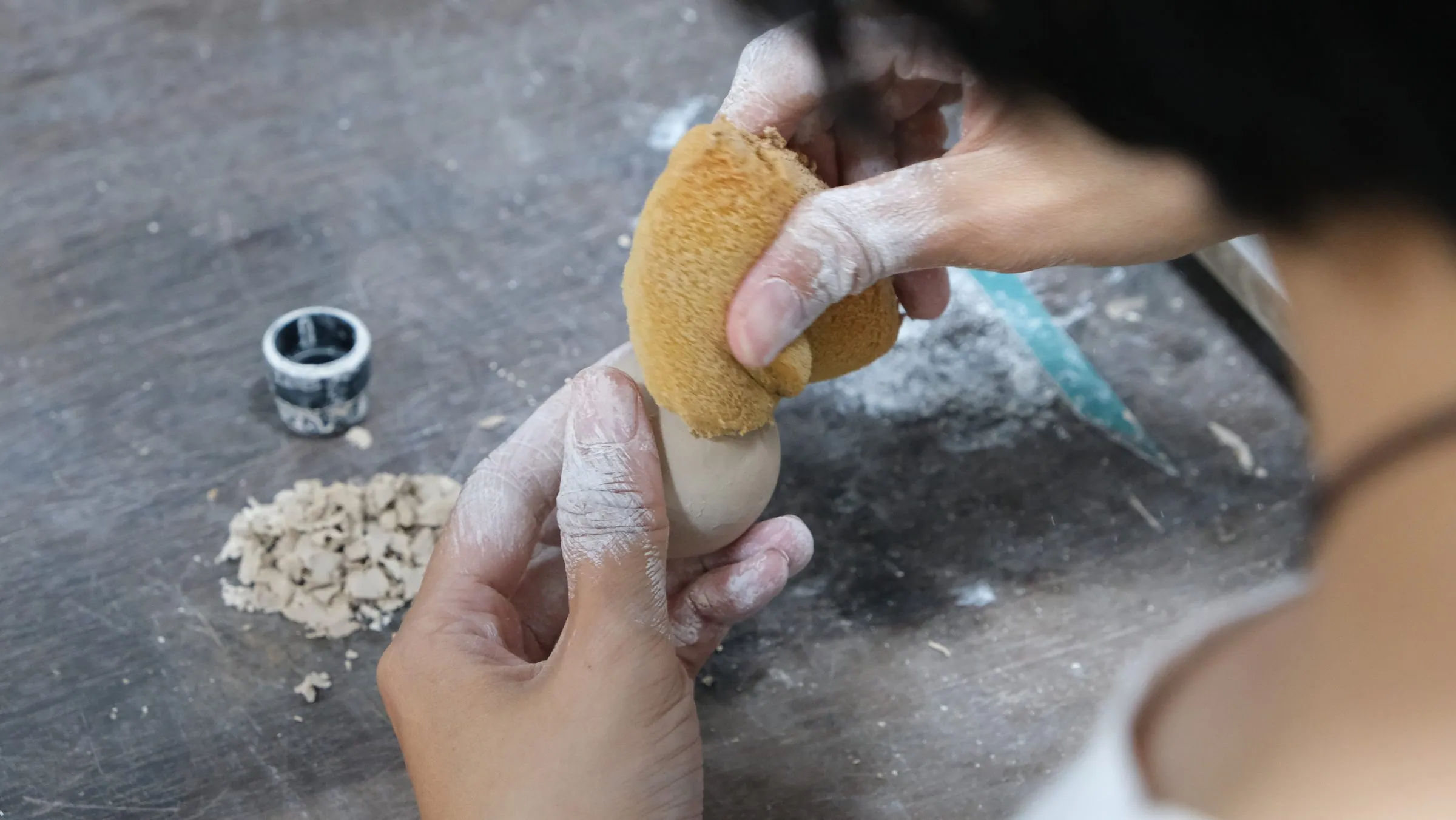 A hand wiping a sphere with a yellow sponge, crumbs of clay and a patterned jar on the table
