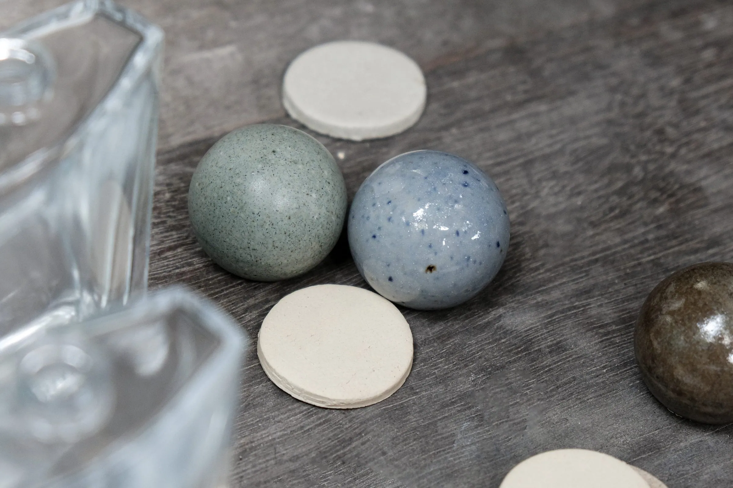 Fired ceramic spheres in blue-grey and speckled glaze beside a round ceramic disc, a glass bottle out of focus in the foreground