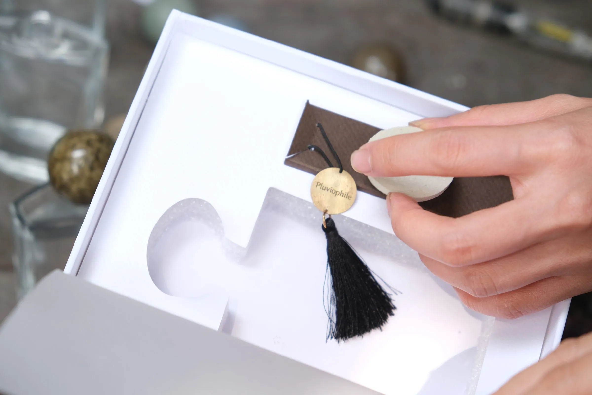 A hand placing a round cream ceramic charm and a black tassel into a white box with shaped hollows