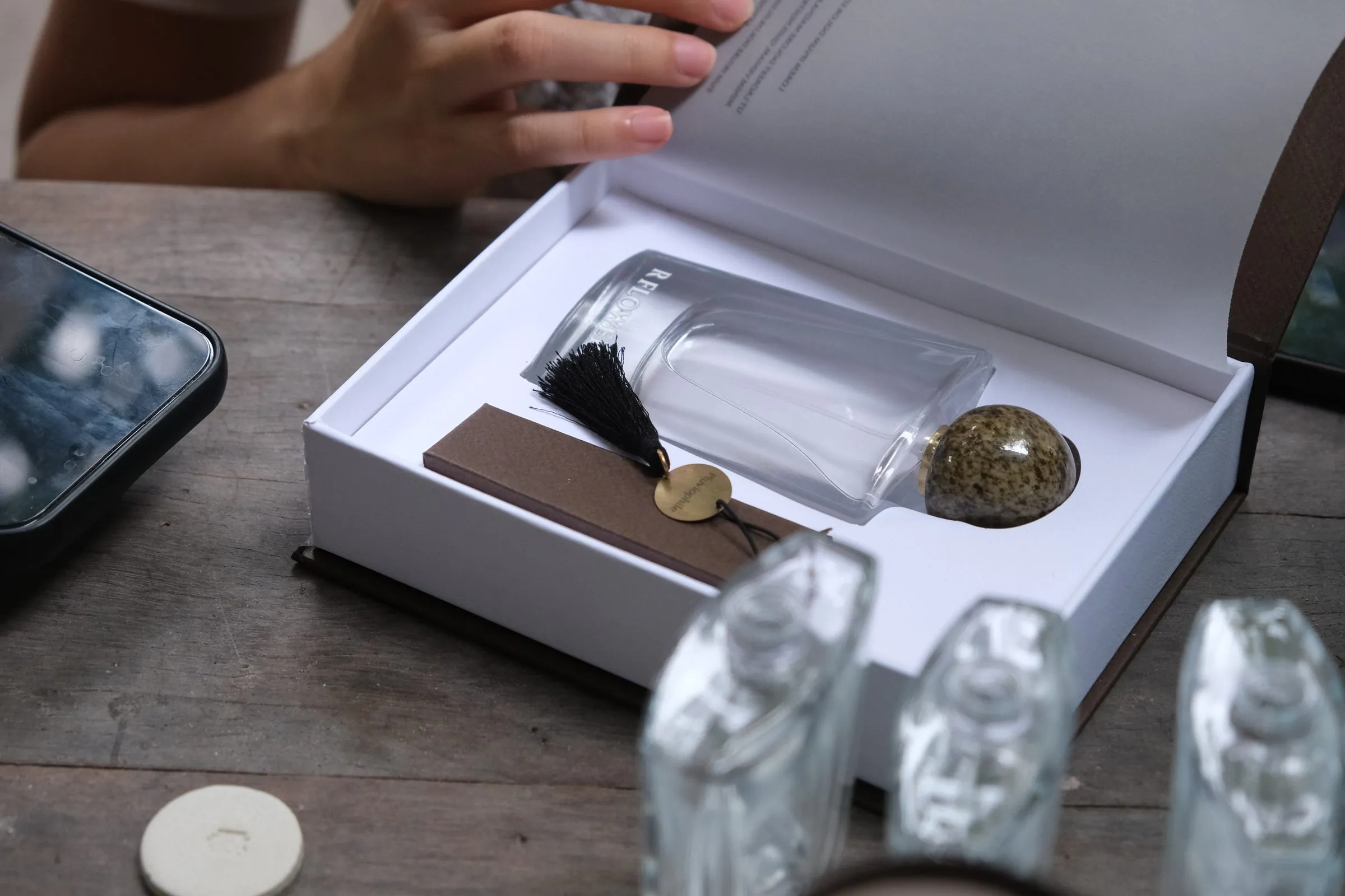 An open white box holding a glass bottle, a black tassel and a brown-glazed ceramic cap; a hand rests on the lid