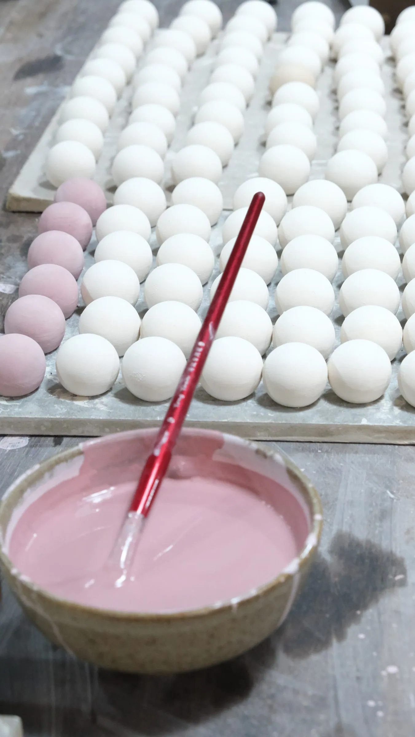 A bowl of pink glaze with a red-handled brush beside a tray of white spheres, the left row already pink