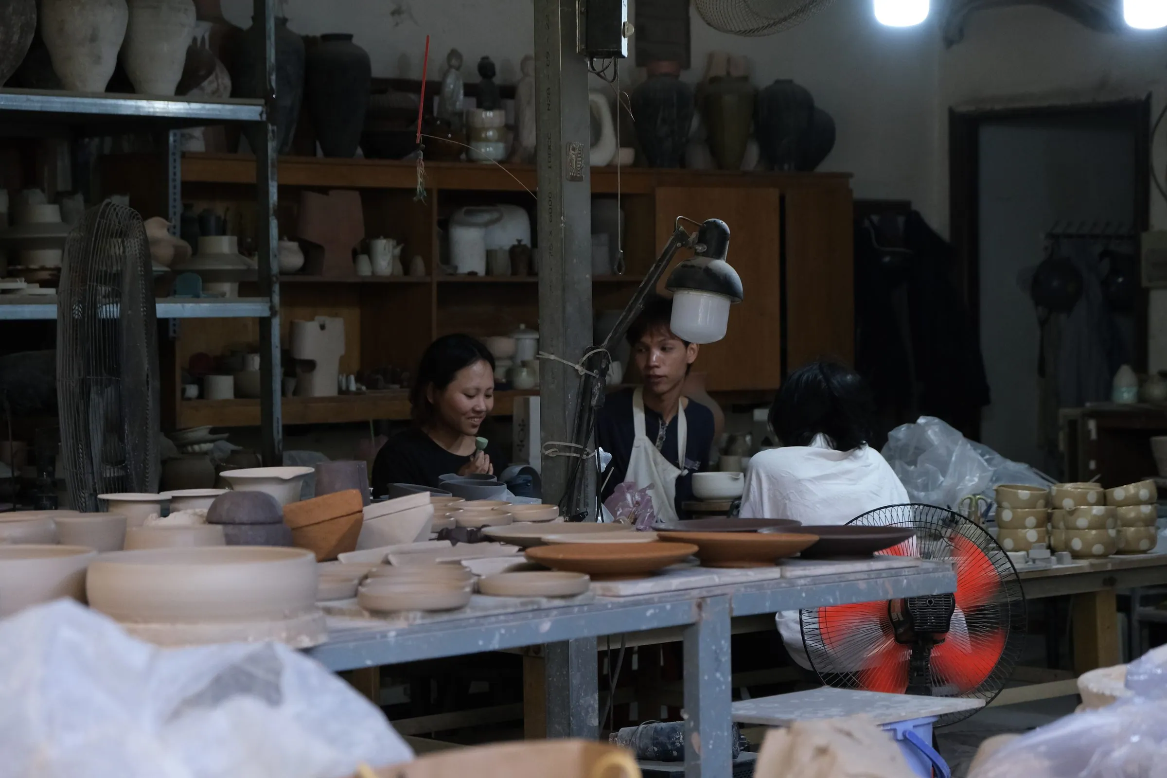 The pottery studio: two people at work at a long table, shelves of pottery behind