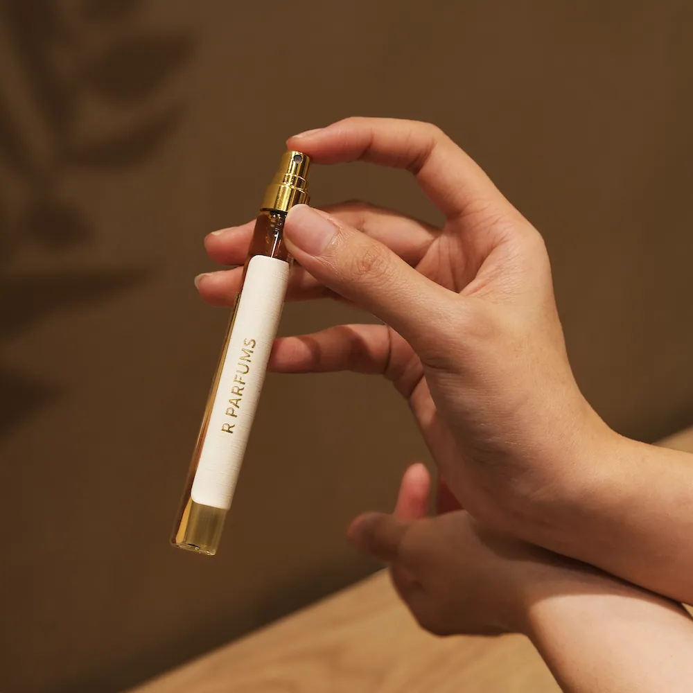 Une main tient le flacon fin de 12 ml, R PARFUMS imprimé sur toute sa hauteur
