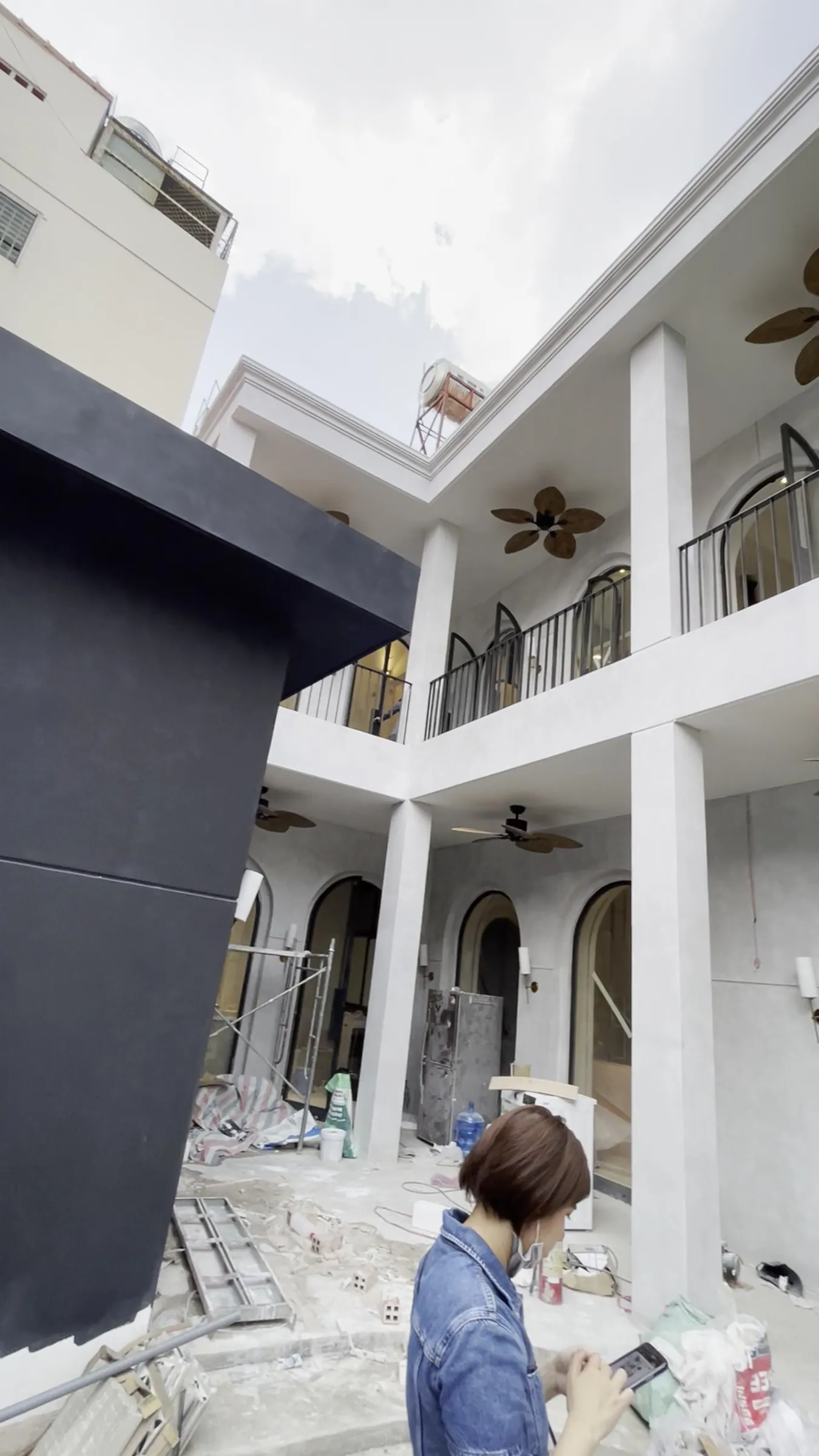 The newly finished two-storey white façade, seen from the courtyard: columns, balconies with iron railings, arched doors, a ceiling fan with leaf-shaped blades; on the left, a block of dark grey wall