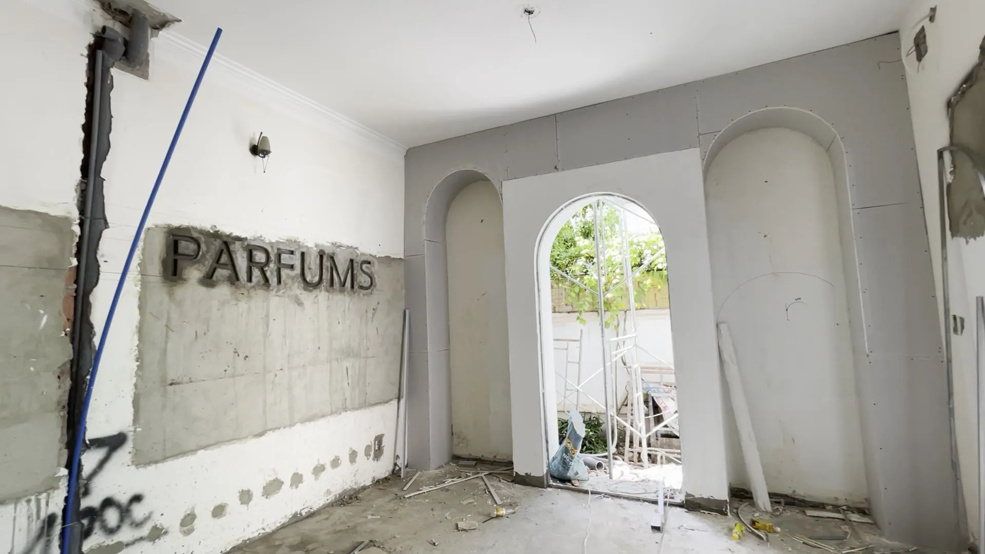 Metal letters spelling PARFUMS on a raw concrete wall; in the middle of the room a white arch opens onto greenery, an arched niche on either side, electrical conduit still running along the wall