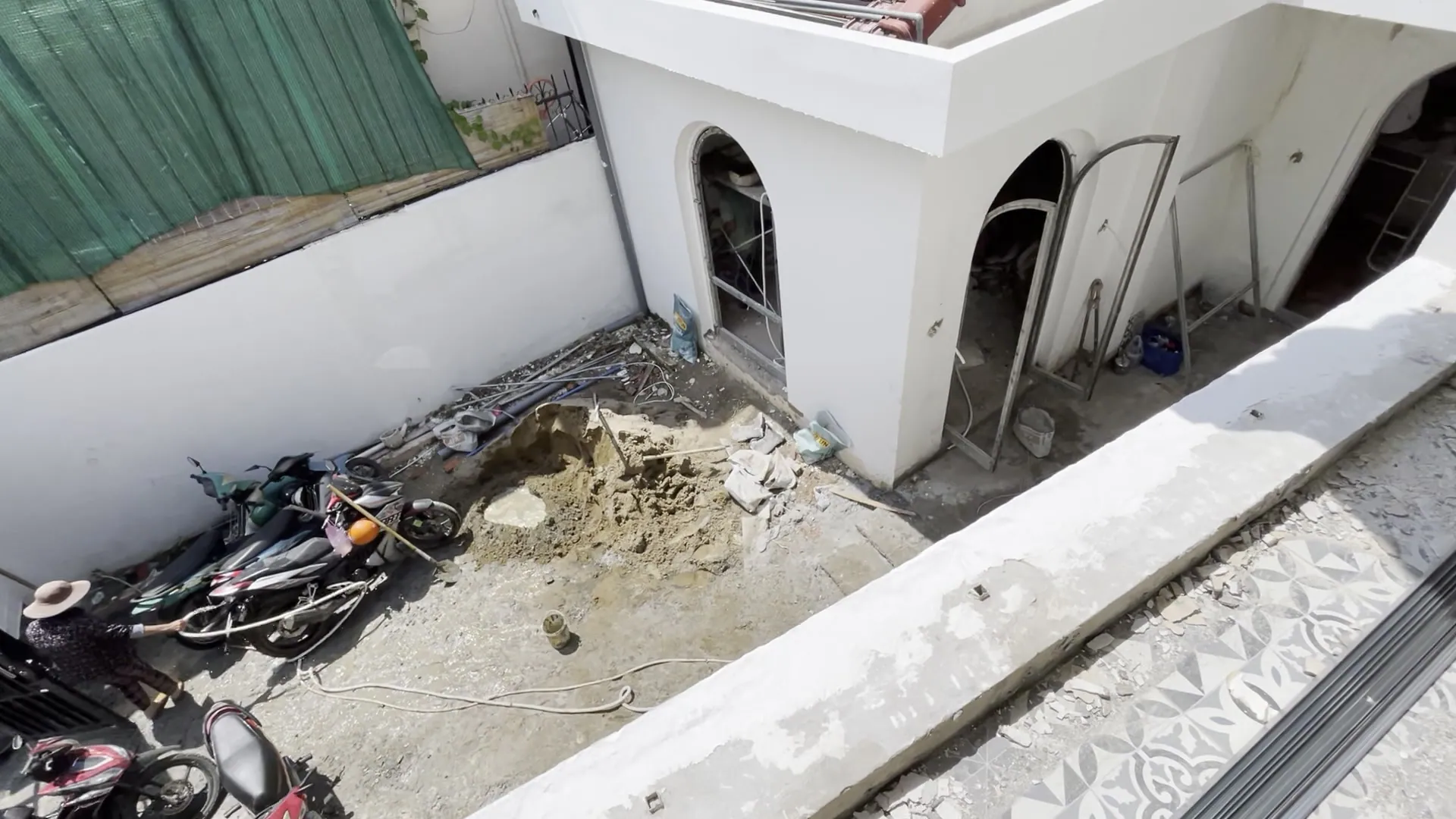 Looking down into the courtyard under construction: a heap of sand, a motorbike, a steel arched door frame not yet glazed, a worker in a hat at the lower corner; along the right edge, the grey patterned tiles of the upper corridor