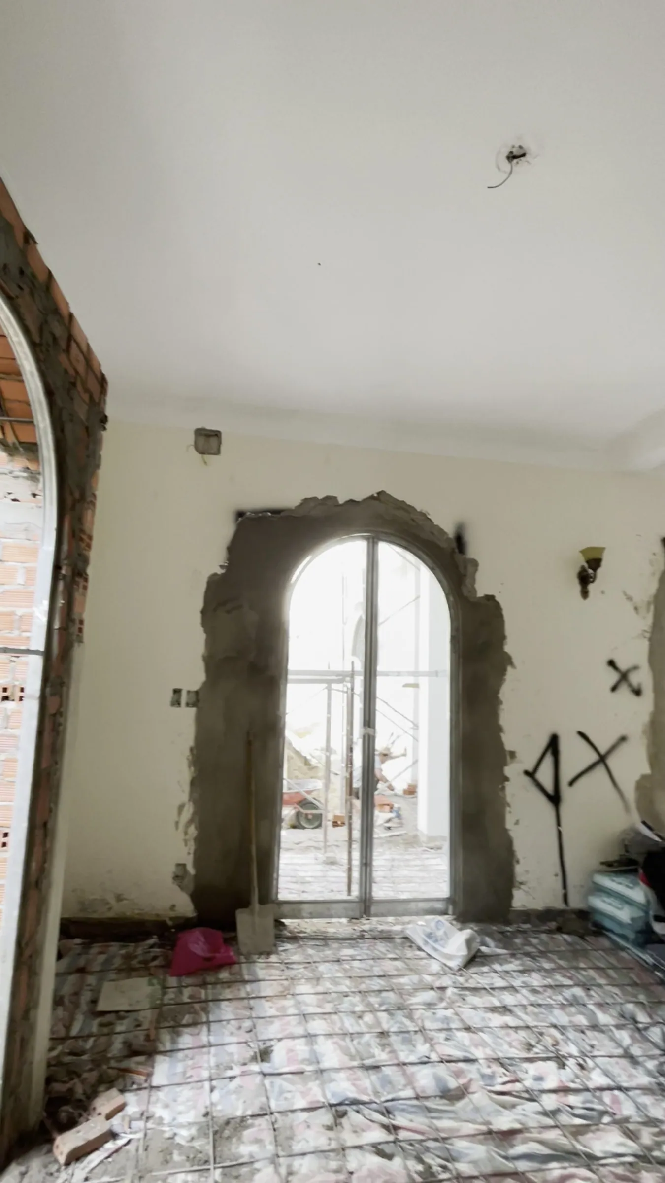 A room under renovation: an arched opening newly widened, its edges raw grey mortar, an arched glass frame standing inside; an X sprayed in paint on the wall, rubble still on the floor
