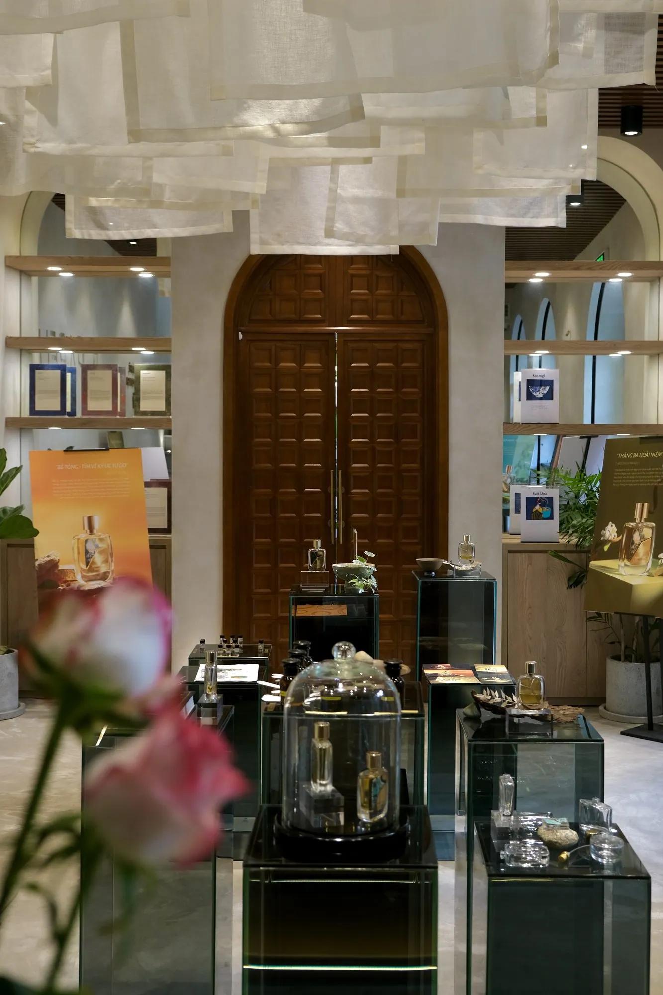 The front room with no one in it: a carved wooden arched door, linen hanging from the ceiling, bottles under glass on dark display stands, an orange story poster for Béton Chaud, and a pink rose out of focus in the foreground