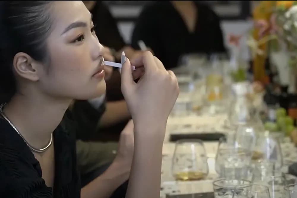 女孩在长桌旁嗅闻试香纸，桌上摆着玻璃杯与花，摄于一场品香聚会。
