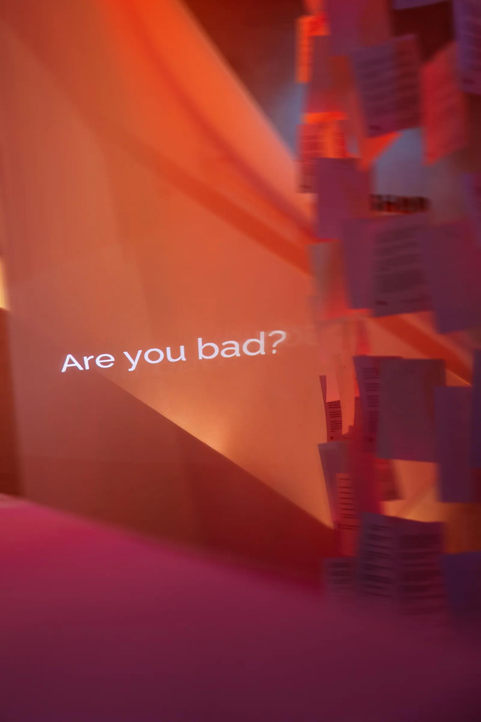 Dòng chữ chiếu «Are you bad?» trên vách cam, bên phải là rừng thư nhoè chuyển động