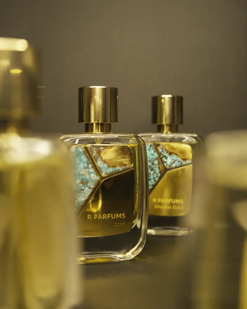 Hai chai R Parfums 221B và Shadow Éclat cạnh nhau, mặt chai ốp tấm đồng thau patina xanh ngọc, nắp vàng