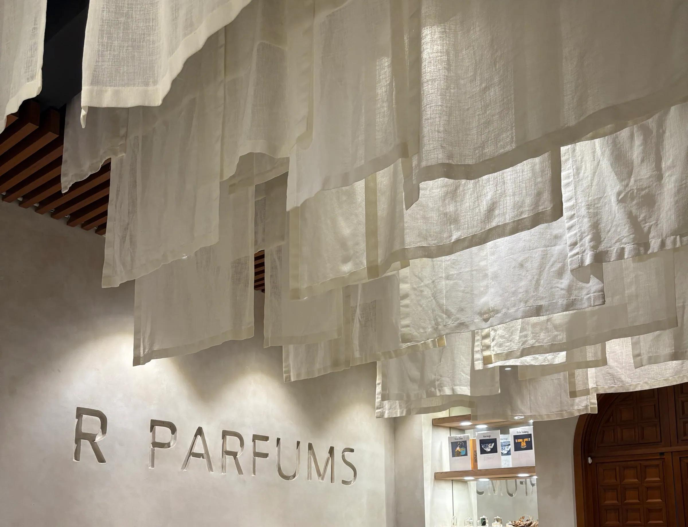 层层亚麻布从天花板垂下，下方浅色灰泥墙上刻着 R PARFUMS 字样，墙边靠着一张长木桌。