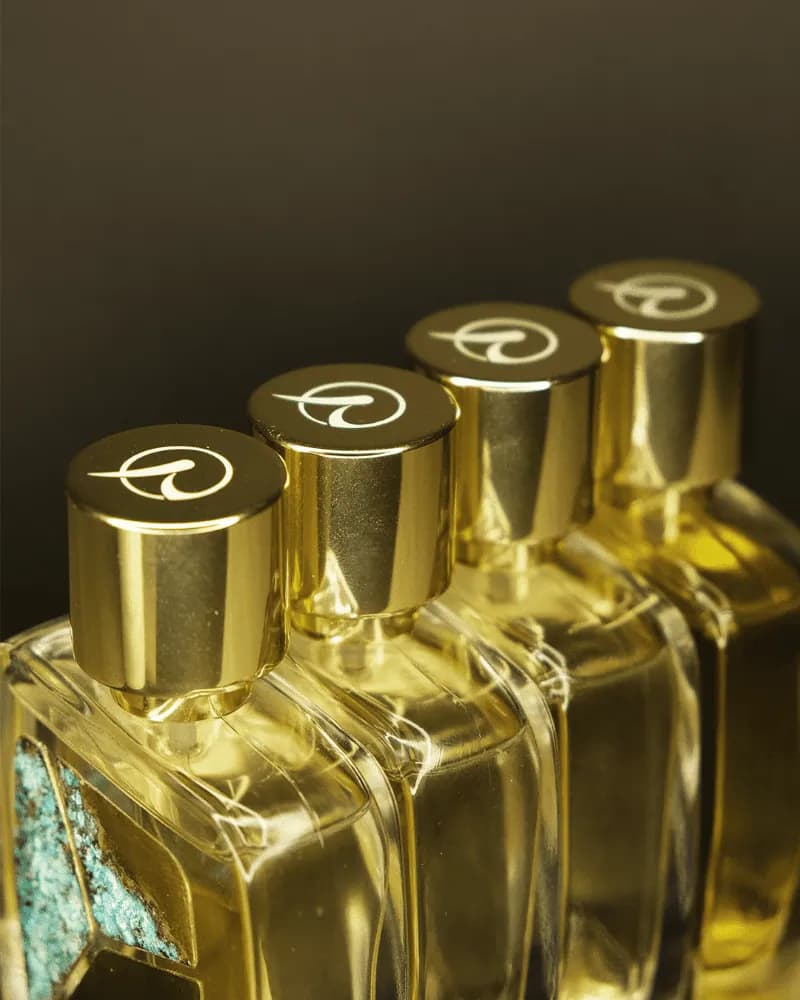 Bốn chai R Parfums xếp hàng chéo, nắp vàng khắc logo chữ R, thấp thoáng tấm patina xanh ngọc trên thân chai