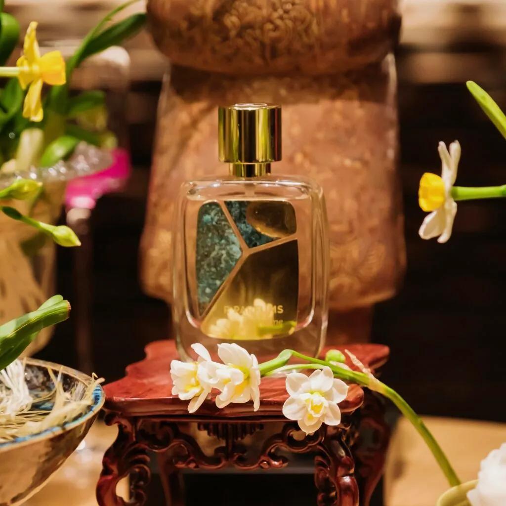 金色瓶盖的 Eau de Hajime 香水瓶,瓶身拼贴泛着铜绿的铜片,立在雕花木座上,四周是白色水仙。