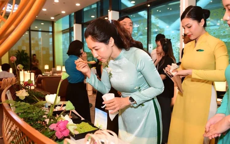 A guest in a pale teal áo dài samples food at a lotus and young-rice display beside an attendant in yellow